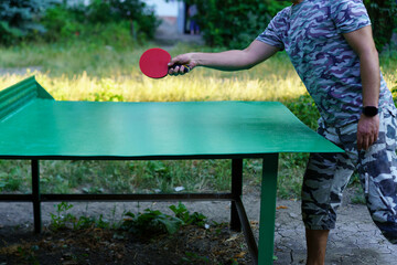 Man playing table tennis in a sunny outdoor garden setting while wearing camouflage clothing