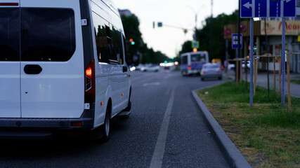 White van driving on a city street in the evening with traffic signals and parked cars lining the road