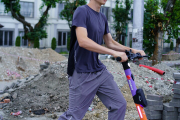 A young adult navigates an electric scooter through a construction site in a city, with visible...