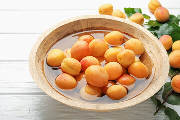 Fresh ripe apricots in bowl with water and green leaves on white wooden table, closeup