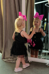 a little girl in a black dress and a pink bow stands by a large mirror and looks into it