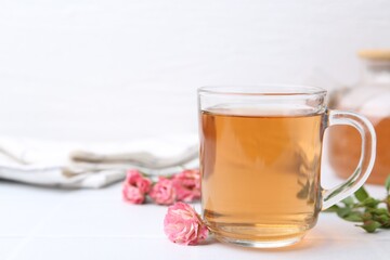 Aromatic tea in glass cup, roses and green leaves on white table, closeup. Space for text