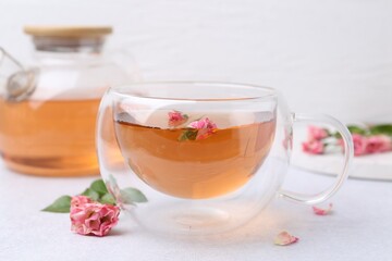 Aromatic tea in glass cup, teapot, roses and green leaves on light table, closeup