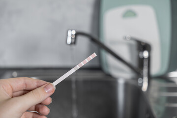 Hand holding a water test strip showing purity results against a kitchen sink and faucet, indicating home water quality assessment
