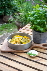 Freshly prepared vegetable dish featuring colorful zucchini, squash, and herbs served in a modern gray bowl on a rustic wooden table with greenery in the background