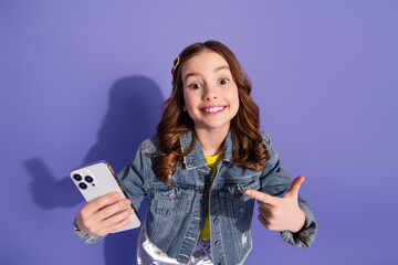 Cute young girl smiling while holding a smartphone and gesturing confidently against a purple...