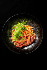 Asian noodle bowl with roasted meat, red bell peppers, cilantro, and sesame seeds served in a black dish on a dark background, top view, close-up.