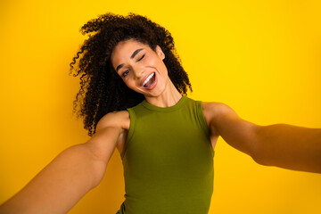 Cheerful young woman with curly hair taking a selfie in a vivid yellow background wearing a trendy...