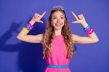 Charming young woman in a stylish pink dress with a crown posing joyfully against a vibrant purple...