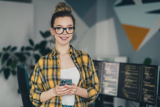Young female programmer holding smartphone near computer monitors, smiling and working in a modern office environment