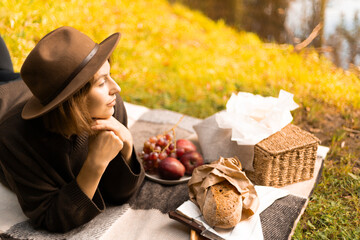 A scenic lakeside picnic in autumn — a woman lies back on a blanket with cozy food, a teacup, and...