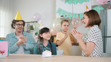 Happy cute child making a wish and blowing birthday cake candle surrounded by family wearing party...