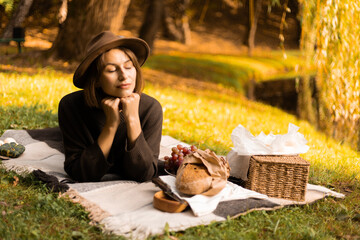 A cozy and romantic lakeside picnic: woman in a wide-brimmed hat rests among apples, croissants,...