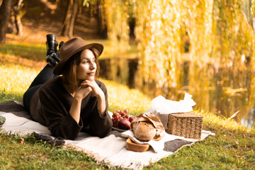 A felt-hatted woman reclines beside a calm lake, indulging in a rustic fall picnic of fruit, tea, and pastries.