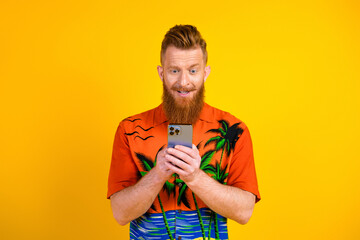 Cheerful man with red hair using smartphone in bright tropical shirt on a vibrant yellow background