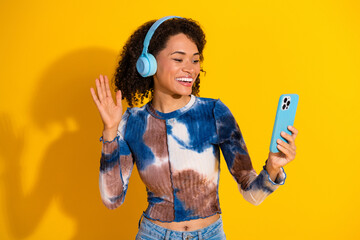 Young woman wearing headphones smiling and waving while interacting on a smartphone against a...