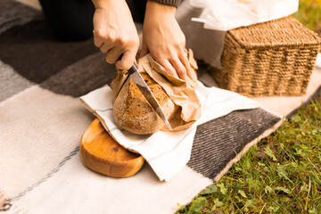 Close-up of woman’s hands cutting bread on a cozy fall picnic setup near the water, surrounded by...
