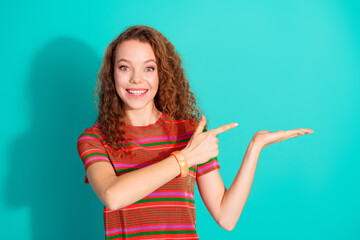 Happy young woman with curly hair pointing at copy space with one hand on a vibrant turquoise...