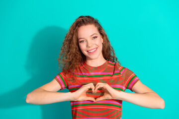 Smiling young woman with curly hair in colorful striped shirt making a heart gesture on a bright...
