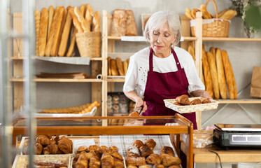 Friendly female baker in apron putting hot delicious croissants to the showcase for sale