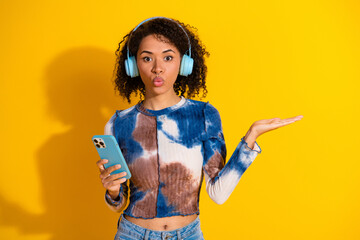Young woman with curly hair listening to music and holding a smartphone against a vibrant yellow...