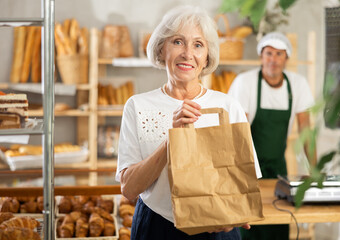 Senior woman bakery customer stands in sales area with paper bag in hands. Client bought fresh...