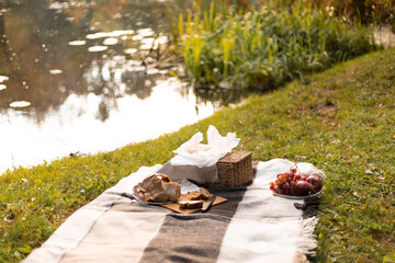 A scenic picnic still life featuring a wicker basket, bread, and fruit on a blanket near a tranquil...
