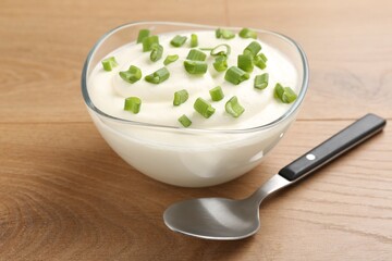 Delicious yogurt in bowl, green onions and spoon on wooden table, closeup