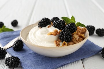 Tasty yogurt with granola and blackberries in bowl on white wooden table, closeup