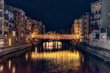 Illuminated Eiffel Bridge Pont des Peixateries Velles in Girona, Spain at night with yellow lighting reflecting in river Onyar. Historic houses line both sides of waterfront in old town quarter