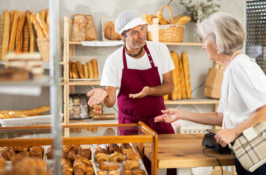 In sales area of bakery near window, customer is talking to owner of bakery. Man seller communicates with mature woman buyer, discusses baking and makes casual jokes