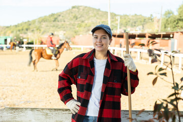 Portrait of happy female stables worker in plaid shirt holding working tool rake or pitchfork...