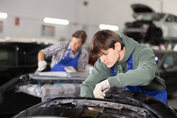 Focused young auto body technician in casual olive hoodie and blue overalls carefully repairing car bumper in professional specialized workshop