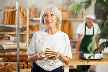 Senior female customer in sales area of bakery store chooses sweets for tea. She takes piece of...