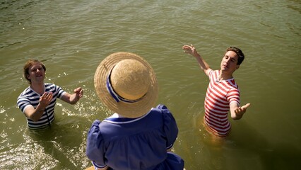 Two men in vintage striped swimsuits are playfully interacting with a woman in a blue dress and...