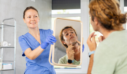 Fototapeta premium Adult man examining his face after cosmetology procedures in mirror reflection in aesthetic medicine office..