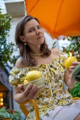 A woman in a floral dress sits outdoors, holding lemons and looking relaxed under an orange umbrella