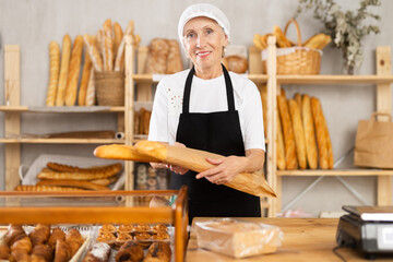 Kind old female marketer offering baguettes in pulp bag in bakehouse