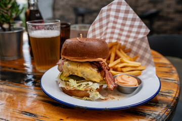 Delicious gourmet bacon cheeseburger with crispy fries and dipping sauce served on rustic wooden table with cold beer in a casual restaurant setting