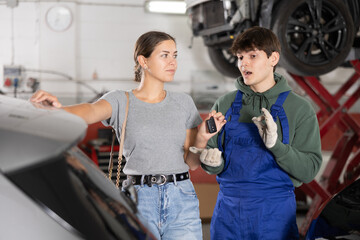 Smiling female client giving car keys to friendly young mechanic in blue overalls, who enthusiastically explaining details of upcoming repair in auto shop..