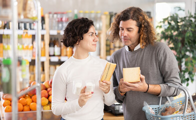Couple choosing compare varieties some sorts of cheese, doubt and think before buying. Client choose and decide purchase cheese delicious package, inspect good.