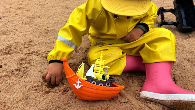 Child in yellow rain suit and pink boots playing with a toy pirate ship in the sand. Close-up of creative outdoor play during rainy weather. - Powered by Adobe