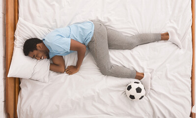 Laughing black sport fan sleeping with soccer ball, dreaming about playing football, top view