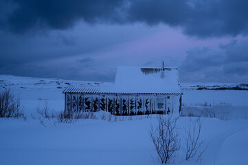 Remote cabin blanketed in snow under a dramatic winter sky, surrounded by a vast snowy landscape and soft twilight hues