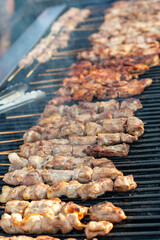A large chargrill with rows of marinated pork, chicken, and lamb chunks on wooden skewers, barbequing. The barbecue-roasted and seared protein has a sauce on the meat. The street food is for sale.