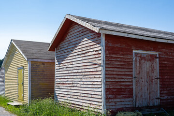 A row of small colorful painted huts or sheds made of clapboard wood. The exterior walls are worn and weathered with paint peeling. The doors are shutter style. The ground is covered with green grass.