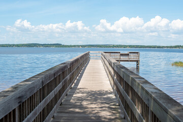 Naklejka premium The grey, worn plank-style pier juts out into a lake on all three sides, with tree-covered land in the background under a blue sky with clouds. There are wide handrails on both sides of the wharf.