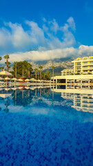A luxurious hotel stands tall, reflected in a tranquil pool beneath blue skies. Majestic mountains...