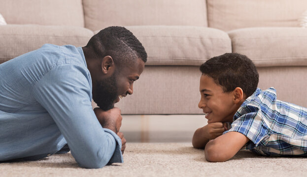 Profile portrait of handsome african american man and his teen son lying on carpet at home and looking at each other