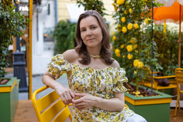 A woman in a white floral dress sits outdoors, surrounded by vibrant yellow roses, enjoying a bright and cheerful day